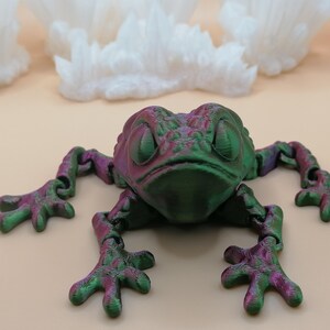 May include: A 3D-printed frog figurine in shades of green and purple. The frog has articulated limbs and a textured surface. The background features white, cloud-like structures against a light beige backdrop.