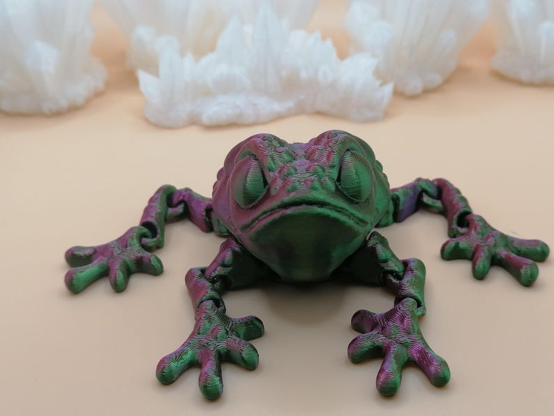 May include: A 3D-printed frog figurine in shades of green and purple. The frog has articulated limbs and a textured surface. The background features white, cloud-like structures against a light beige backdrop.