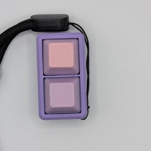 May include: A two-button keycap set in a lavender frame, with a black lanyard. The top button is pink, and the bottom button is lavender with diagonal lines. The keycaps are square and have a smooth surface.