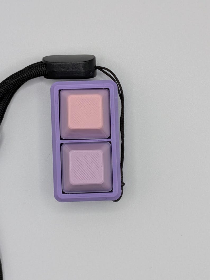 May include: A two-button keycap set in a lavender frame, with a black lanyard. The top button is pink, and the bottom button is lavender with diagonal lines. The keycaps are square and have a smooth surface.
