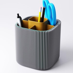 May include: A grey and yellow desk tidy with a ribbed exterior. The organiser contains a black pen, a yellow pencil, a black pencil, and blue scissors. The interior is divided into compartments for stationery storage.