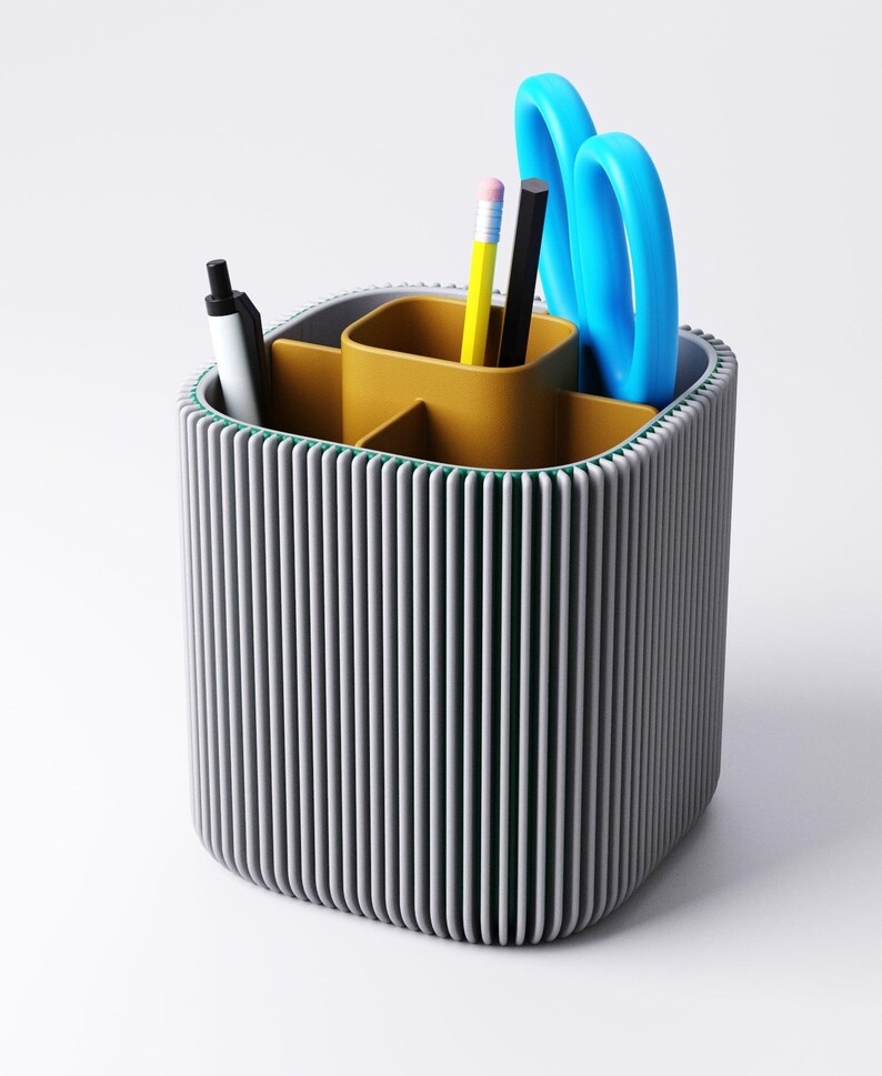 May include: A grey and yellow desk tidy with a ribbed exterior. The organiser contains a black pen, a yellow pencil, a black pencil, and blue scissors. The interior is divided into compartments for stationery storage.