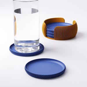 May include: A set of blue and orange coasters. The coasters are round with a ribbed edge. A glass of water sits on one coaster. The set includes a holder with a stack of coasters.