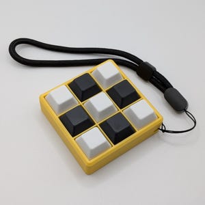 May include: A yellow square device with nine black and white square buttons, resembling a miniature keyboard, is attached to a black lanyard. The buttons are arranged in a 3x3 grid. The device is set against a plain white background.