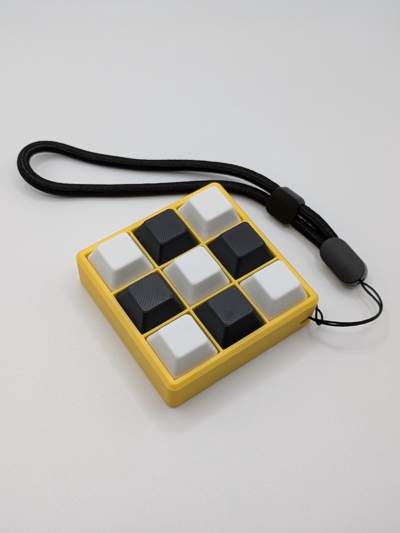 May include: A yellow square device with nine black and white square buttons, resembling a miniature keyboard, is attached to a black lanyard. The buttons are arranged in a 3x3 grid. The device is set against a plain white background.