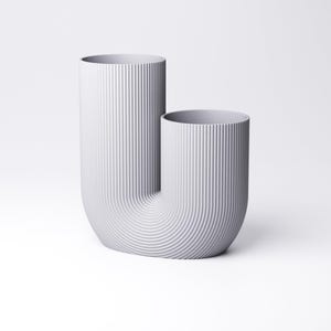 K&ouml;nnte beinhalten: Eine hellgraue U-f&ouml;rmige Vase mit einer vertikal gerippten Textur auf der Au&szlig;enseite. Das Objekt besteht aus zwei zylindrischen &Ouml;ffnungen unterschiedlicher H&ouml;he, die durch eine geschwungene Basis verbunden sind und eine durchgehende Oberfl&auml;che bilden.