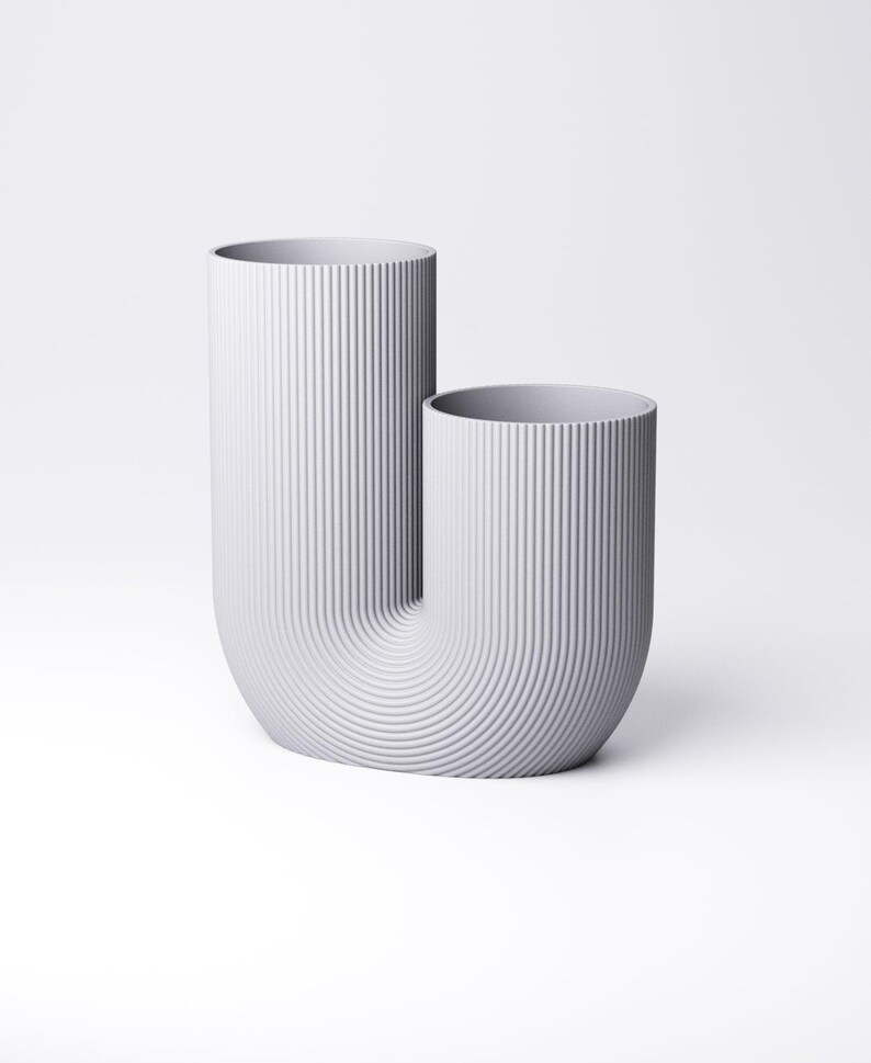 K&ouml;nnte beinhalten: Eine hellgraue U-f&ouml;rmige Vase mit einer vertikal gerippten Textur auf der Au&szlig;enseite. Das Objekt besteht aus zwei zylindrischen &Ouml;ffnungen unterschiedlicher H&ouml;he, die durch eine geschwungene Basis verbunden sind und eine durchgehende Oberfl&auml;che bilden.