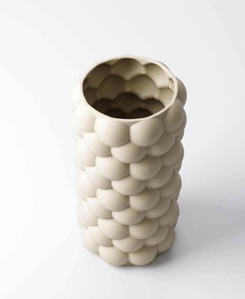 May include: A cream-colored, cylindrical vase with a textured surface. The exterior is covered in a pattern of rounded, bubble-like shapes. The vase has a wide opening at the top and is set against a white background. A decorative home decor item.