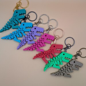 May include: A collection of colorful dinosaur keychains in shades of teal, blue, purple, pink, red, green, and gray. Each articulated dinosaur is attached to a metal keyring with a lobster clasp. The keychains are arranged in a diagonal row.