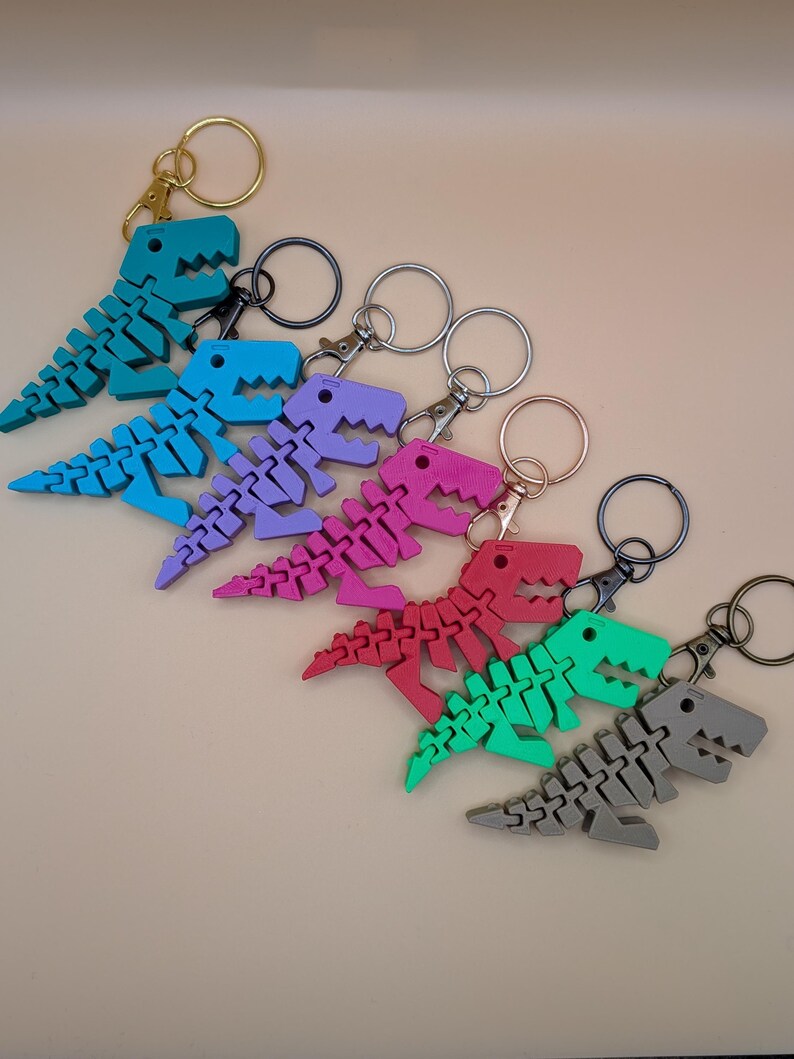 May include: A collection of colorful dinosaur keychains in shades of teal, blue, purple, pink, red, green, and gray. Each articulated dinosaur is attached to a metal keyring with a lobster clasp. The keychains are arranged in a diagonal row.
