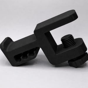 Puede incluir: Una abrazadera negra en forma de C con un tornillo roscado y una perilla texturizada. La abrazadera tiene un cuerpo rectangular con un extremo curvo y tres agujeros circulares. El objeto est&aacute; hecho de material negro mate.