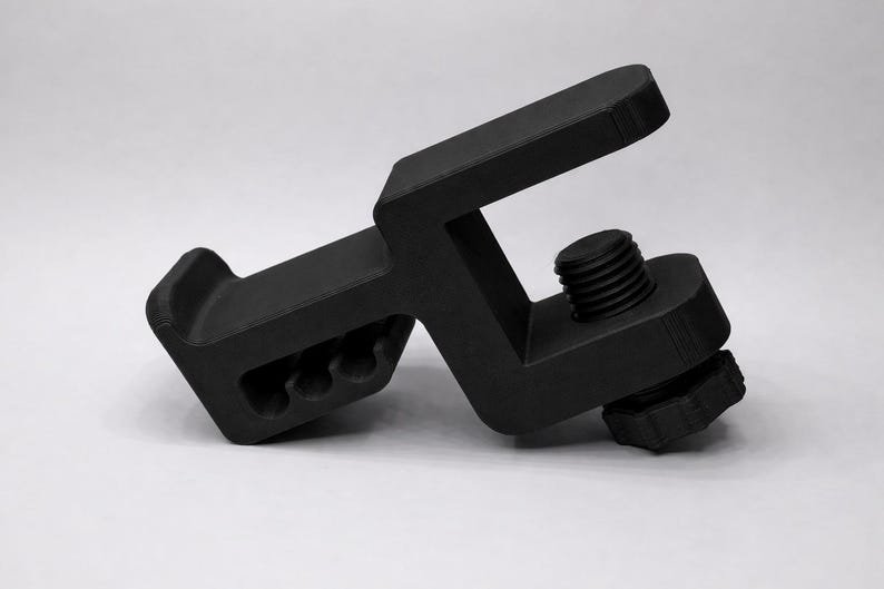 Puede incluir: Una abrazadera negra en forma de C con un tornillo roscado y una perilla texturizada. La abrazadera tiene un cuerpo rectangular con un extremo curvo y tres agujeros circulares. El objeto est&aacute; hecho de material negro mate.