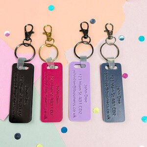 May include: Four keychains in black, gold, black, and silver. Each has a rectangular tag with the name "John Doe", address, and email. The keychains are in black, fuchsia, lavender, and slate blue.