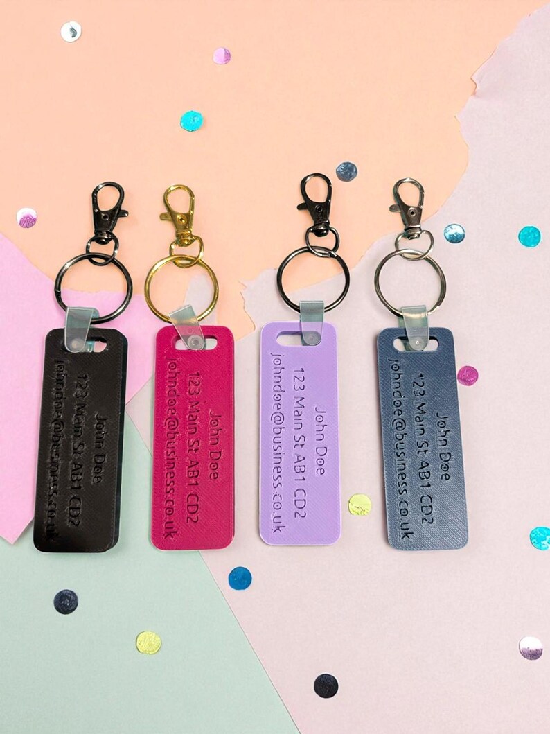 May include: Four keychains in black, gold, black, and silver. Each has a rectangular tag with the name "John Doe", address, and email. The keychains are in black, fuchsia, lavender, and slate blue.