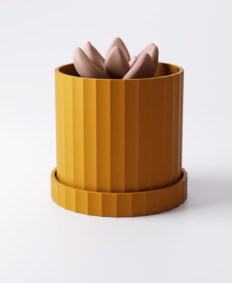 Puede incluir: Una maceta cil&iacute;ndrica de color amarillo mostaza con textura acanalada vertical y una base de plato circular a juego. Dentro de la maceta hay una suculenta de acabado mate suave con p&eacute;talos redondeados de color rosa p&aacute;lido en roseta.