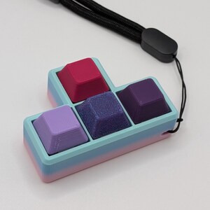 May include: A colorful keyboard keycap set in a Tetris-like shape. The keycaps are various shades of purple, pink, and blue, with a glittery center keycap. It includes a black lanyard for carrying.