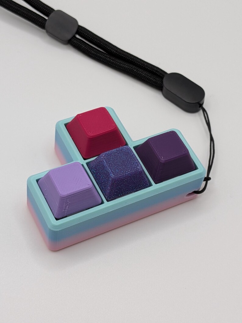 May include: A colorful keyboard keycap set in a Tetris-like shape. The keycaps are various shades of purple, pink, and blue, with a glittery center keycap. It includes a black lanyard for carrying.