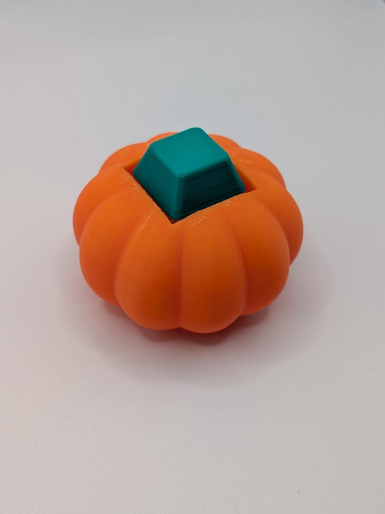 May include: A 3D-printed orange pumpkin with a teal square on top. The pumpkin has a ribbed texture and a hollowed-out center. The teal square is a solid color and sits inside the pumpkin.