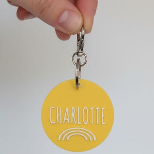 May include: Yellow round keychain with the name "CHARLOTTE" in white lettering and a white rainbow design. The keychain is attached to a silver clasp and key ring.