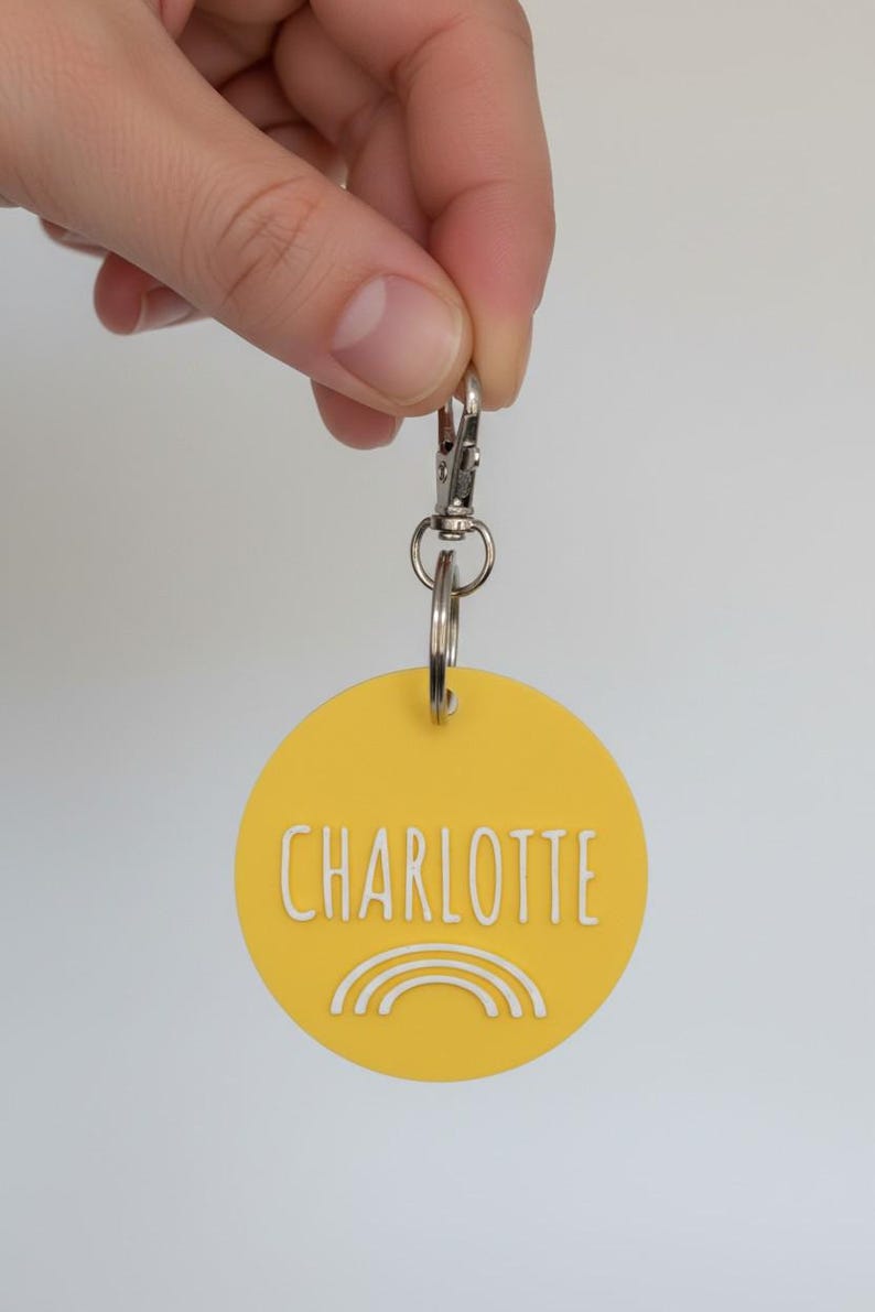 May include: Yellow round keychain with the name "CHARLOTTE" in white lettering and a white rainbow design. The keychain is attached to a silver clasp and key ring.