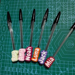 May include: A collection of clear ballpoint pens with black ink, each adorned with a colorful name tag. The tags display names like "Amy", "Noah", "Luna", "Jack", and "Mia" in bold, white lettering. The pens rest on a green cutting mat.