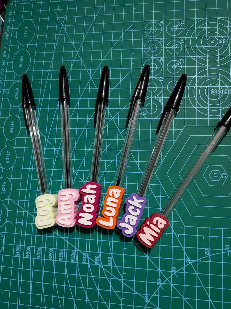 May include: A collection of clear ballpoint pens with black ink, each adorned with a colorful name tag. The tags display names like "Amy", "Noah", "Luna", "Jack", and "Mia" in bold, white lettering. The pens rest on a green cutting mat.