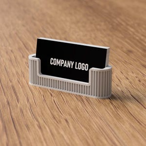 May include: A business card holder with a black card that reads "COMPANY LOGO" in white. The holder is light gray and has a ribbed design. The background is a wooden surface.