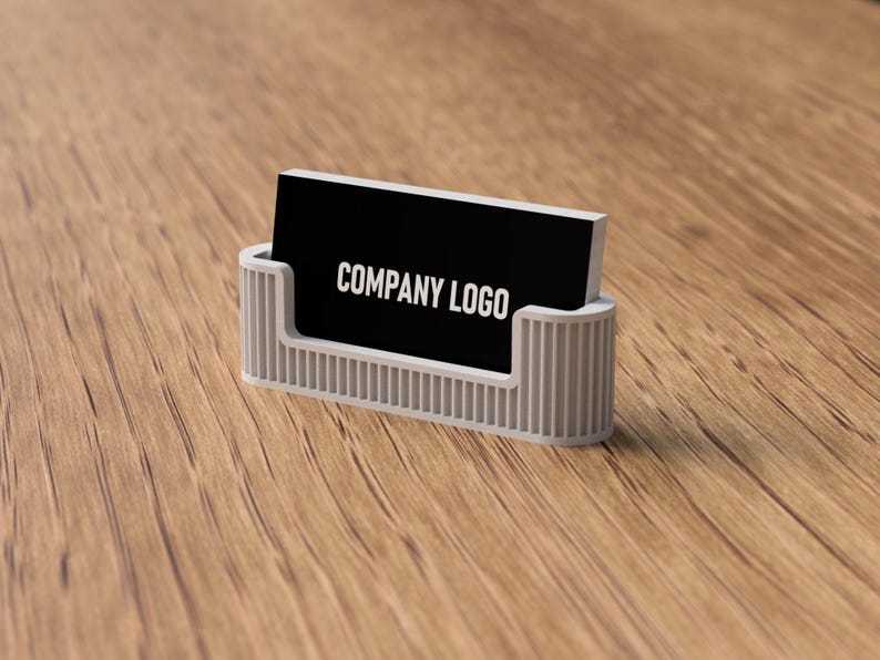 May include: A business card holder with a black card that reads "COMPANY LOGO" in white. The holder is light gray and has a ribbed design. The background is a wooden surface.