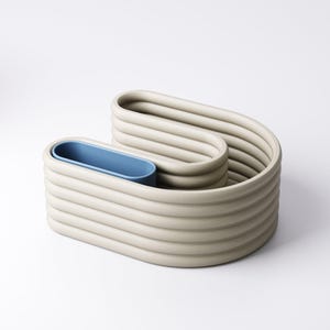 May include: An abstract sculpture in a neutral beige colour with a blue interior. The piece has a unique, wavy design with a series of horizontal, rounded ridges. The blue interior provides a striking contrast to the beige exterior. The sculpture is on a white background.