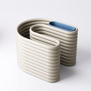 May include: An abstract sculpture in a neutral beige colour. The piece has a unique, wavy design with horizontal ridges and a blue, oval-shaped insert. The sculpture is likely made of ceramic or a similar material.
