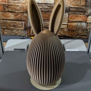 Puede incluir: Figura decorativa de conejo de Pascua marr&oacute;n. El conejo tiene un cuerpo en forma de huevo con rayas verticales y orejas altas. La figura est&aacute; hecha de un material marr&oacute;n claro y se apoya en una base redonda de color claro.