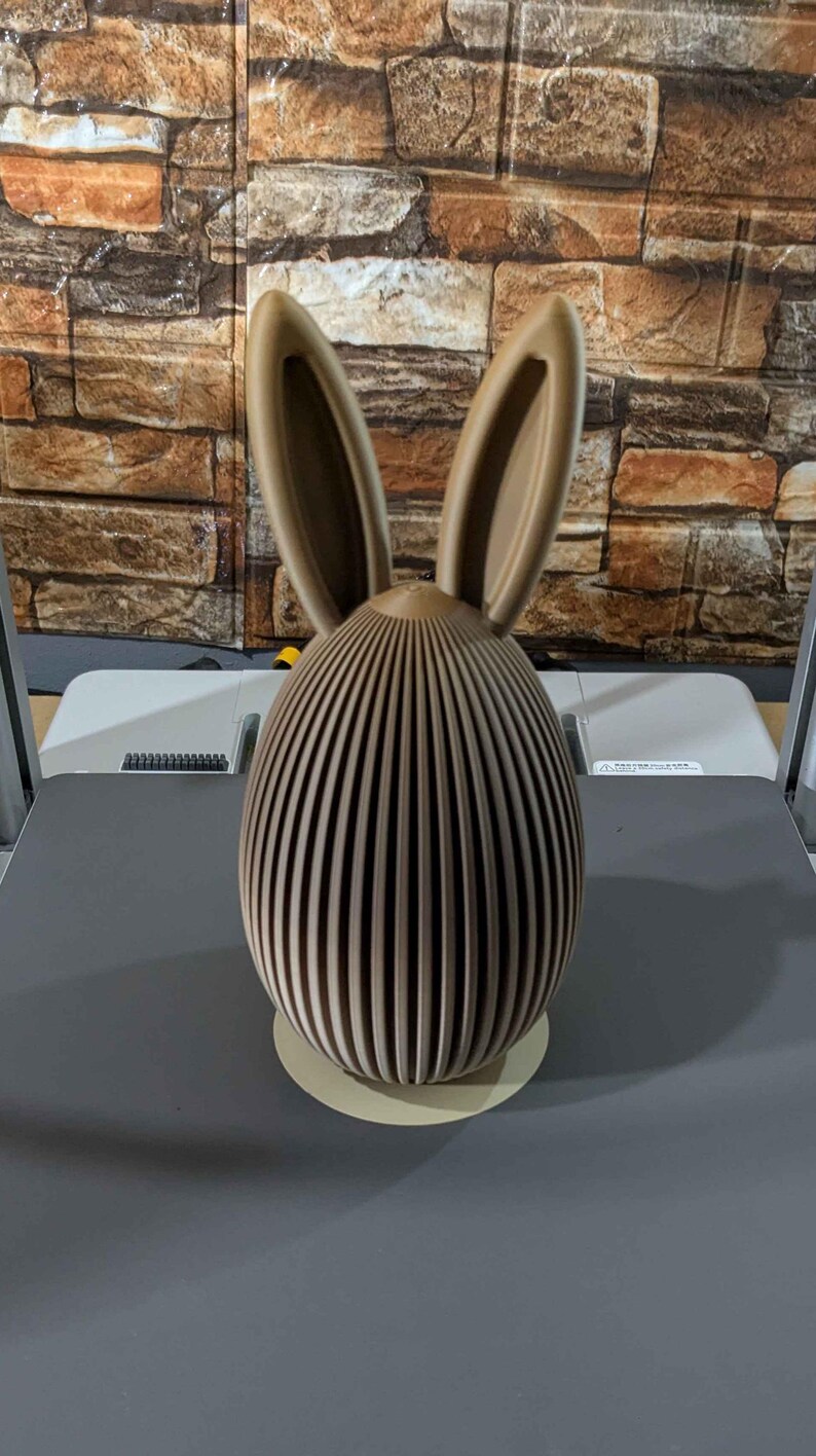 Puede incluir: Figura decorativa de conejo de Pascua marr&oacute;n. El conejo tiene un cuerpo en forma de huevo con rayas verticales y orejas altas. La figura est&aacute; hecha de un material marr&oacute;n claro y se apoya en una base redonda de color claro.
