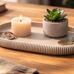 Puede incluir: Una bandeja ovalada beige con borde acanalado contiene una vela encendida, una peque&ntilde;a planta suculenta en maceta, monedas y anillos. La bandeja est&aacute; sobre una superficie de madera, creando una est&eacute;tica minimalista.