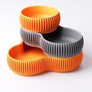 Pu&ograve; includere: Un set di ciotole per animali domestici a tre livelli in arancione e grigio. Le ciotole hanno un design a coste e una forma curva e interconnessa. La ciotola superiore &egrave; arancione, la ciotola centrale &egrave; grigia e la ciotola inferiore &egrave; arancione.