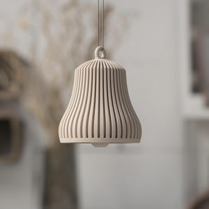 May include: A beige, bell-shaped pendant light with vertical, slotted design. The light hangs from a thin cord and features a rounded top with a small loop. The lamp is made of a light-colored material and is suspended in front of a blurred background.