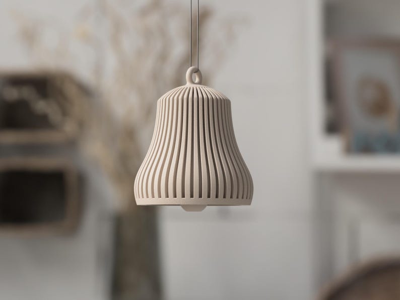 May include: A beige, bell-shaped pendant light with vertical, slotted design. The light hangs from a thin cord and features a rounded top with a small loop. The lamp is made of a light-colored material and is suspended in front of a blurred background.