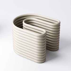 May include: A contemporary sculpture in a pale beige hue. The artwork features two linked, curved rectangular shapes, each with horizontal, ridged detailing. The design is minimalist, with a smooth, matte surface. The sculpture is suitable for interior decor.