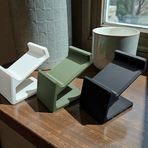 May include: Three minimalist phone stands in white, olive green, and black. Each stand has a simple, angled design with a flat base and a curved top to hold a phone. The stands are displayed on a wooden surface.