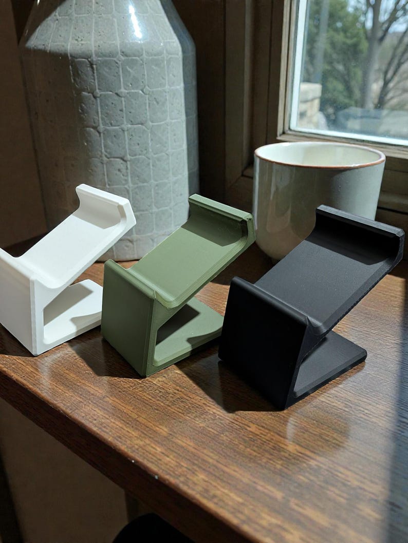 May include: Three minimalist phone stands in white, olive green, and black. Each stand has a simple, angled design with a flat base and a curved top to hold a phone. The stands are displayed on a wooden surface.