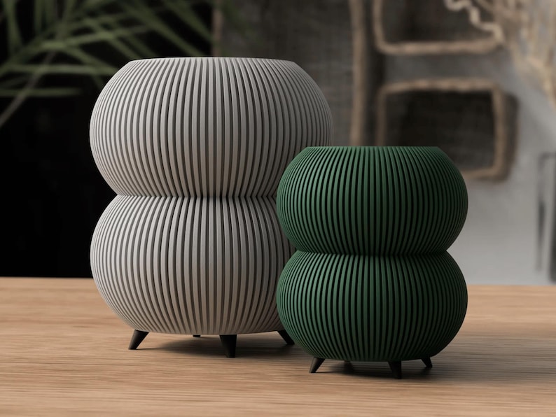 May include: Two decorative, stacked, spherical planters. One is light gray, the other is dark green. Both have vertical, ridged detailing and sit on three small black legs. They are displayed on a light wood surface.