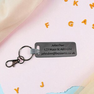 May include: A gray rectangular keychain with a metal clasp and ring. The keychain has the name "John Doe", address, and email address printed on it. Orange letters are scattered around the keychain on a pink surface.