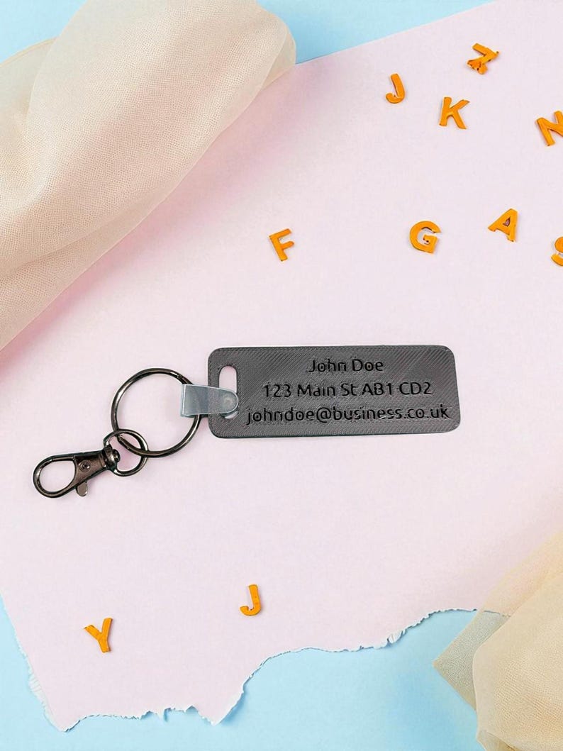 May include: A gray rectangular keychain with a metal clasp and ring. The keychain has the name "John Doe", address, and email address printed on it. Orange letters are scattered around the keychain on a pink surface.