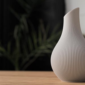 May include: A light gray vase with a textured, ribbed design. The vase has a unique, asymmetrical shape with a slanted top. It sits on a wooden surface, with a blurred background of greenery and a dark wall.