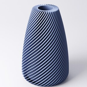 K&ouml;nnte beinhalten: Eine blaue Vase mit einem Spiraldesign. Die Vase hat einen breiten Boden, der sich zu einer schmaleren Oberseite mit einer kreisf&ouml;rmigen &Ouml;ffnung verj&uuml;ngt. Die Au&szlig;enseite weist eine Reihe paralleler, gebogener Linien auf, die eine strukturierte Oberfl&auml;che erzeugen. Die Vase besteht aus einem festen Material.