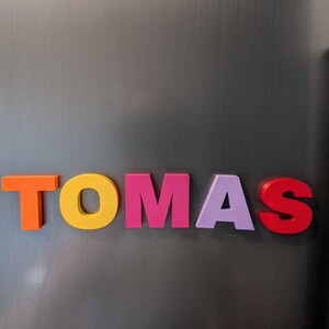 May include: Colorful magnetic letters spelling the name "TOMAS" are arranged on a gray metallic surface. The letters are in various colors: orange, yellow, pink, lavender, and red.