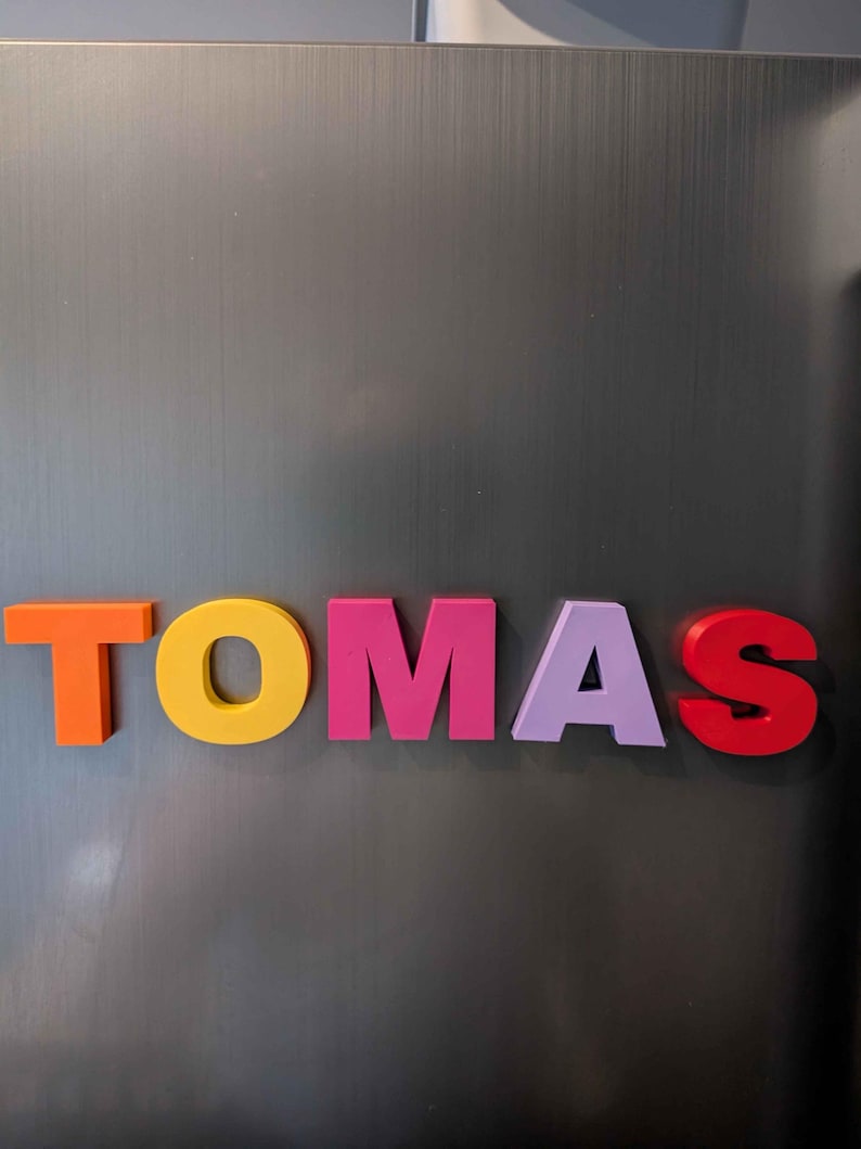 May include: Colorful magnetic letters spelling the name "TOMAS" are arranged on a gray metallic surface. The letters are in various colors: orange, yellow, pink, lavender, and red.