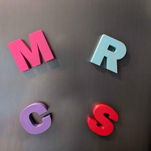 May include: Four colorful magnetic letters: a pink M, a light blue R, a purple C, and a red S. The letters are arranged on a dark gray metallic surface, likely a refrigerator or magnetic board.