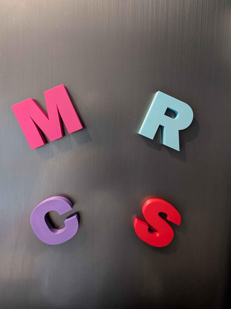 May include: Four colorful magnetic letters: a pink M, a light blue R, a purple C, and a red S. The letters are arranged on a dark gray metallic surface, likely a refrigerator or magnetic board.