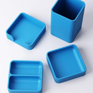 May include: Four bright blue storage containers. The set includes a tall, square pencil holder with vertical ridged sides, two square trays, and a divided tray. The containers are made of a solid, smooth material.