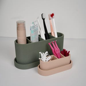 May include: An olive green and a light pink oval bathroom organiser set with vertical ribbed textures. The larger olive green container holds a beige pump bottle, a tube of toothpaste, and four toothbrushes. The smaller pink container holds white dental floss picks and several bright pink interdental brushes.